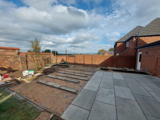 A partially paved outdoor area with brick walls and a house in the background.